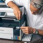 Primer plano de un ingeniero experto de Tecnocom Valle utilizando un destornillador de precisión para realizar el desmontaje y mantenimiento correctivo del sistema de tinta continua en una impresora multifuncional Epson EcoTank. La imagen muestra el proceso de reparación profesional en taller, con herramientas especializadas y hojas de prueba de color sobre el escritorio, ilustrando el servicio de soporte técnico para impresoras de inyección y láser disponible para empresas en Guadalajara de Buga, Cali, Tuluá y el Valle del Cauca.