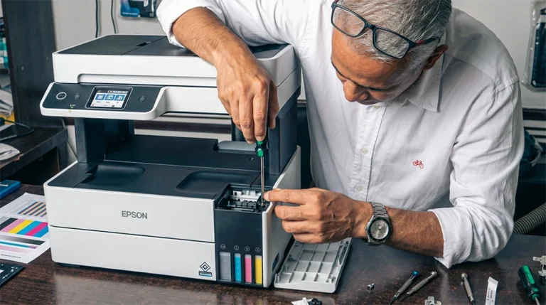 Primer plano de un ingeniero experto de Tecnocom Valle utilizando un destornillador de precisión para realizar el desmontaje y mantenimiento correctivo del sistema de tinta continua en una impresora multifuncional Epson EcoTank. La imagen muestra el proceso de reparación profesional en taller, con herramientas especializadas y hojas de prueba de color sobre el escritorio, ilustrando el servicio de soporte técnico para impresoras de inyección y láser disponible para empresas en Guadalajara de Buga, Cali, Tuluá y el Valle del Cauca.