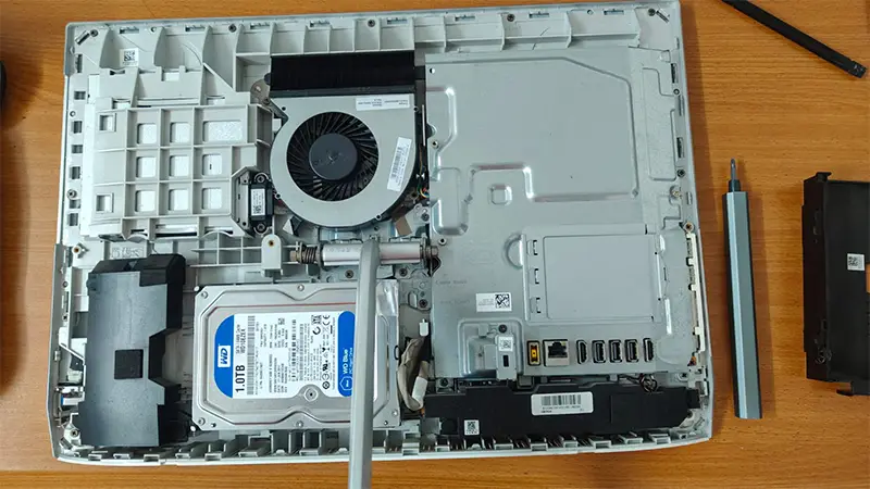 Primer plano del interior desarmado de un computador de escritorio Lenovo Todo en Uno (All-in-One) sobre la mesa de trabajo del servicio técnico de Tecnocom Valle. Se observa el componente causante de la lentitud: un disco duro mecánico original marca WD Blue de 1.0TB de 3.5 pulgadas, ubicado junto al ventilador de refrigeración tipo turbina y el blindaje metálico de la placa base. En la imagen también destaca un destornillador de precisión eléctrico, ilustrando el delicado proceso de mantenimiento y desarme preventivo para clientes en Cali, Buga, Tulúa y el Valle del Cauca.
